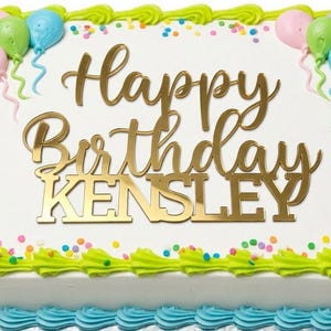 May include: A rectangular birthday cake with white frosting, decorated with green and blue frosting, colorful sprinkles, and gold lettering that reads "Happy Birthday Kensley." Pink, green, and blue balloon decorations are also present.