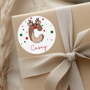 May include: A round white sticker with a reindeer design and the name "Casey" in red script. The reindeer is brown with a red nose and antlers. Red and green dots surround the design. The sticker is on a wrapped gift with a beige ribbon.