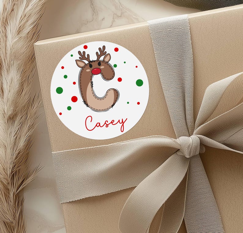 May include: A round white sticker with a reindeer design and the name "Casey" in red script. The reindeer is brown with a red nose and antlers. Red and green dots surround the design. The sticker is on a wrapped gift with a beige ribbon.