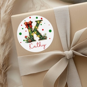 May include: A round sticker with a Christmas theme, featuring the letter "K" decorated with garland and lights. A red bow and the name "Kathy" are also present. Red and green polka dots surround the design. The sticker is on a gift box with a ribbon.