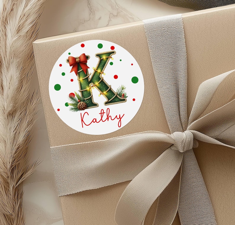 May include: A round sticker with a Christmas theme, featuring the letter "K" decorated with garland and lights. A red bow and the name "Kathy" are also present. Red and green polka dots surround the design. The sticker is on a gift box with a ribbon.
