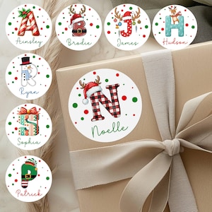 May include: A collection of personalized Christmas gift stickers. Each sticker features a letter of the alphabet, a name, and festive holiday designs like reindeer antlers, Santa hats, and Christmas presents. The stickers are round with red and green accents.