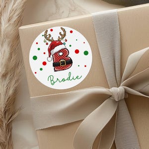 May include: A round white sticker with a Christmas theme, featuring the letter "B" in a Santa Claus outfit with reindeer antlers. The name "Brodie" is written in green script below the letter. Red and green dots surround the sticker.