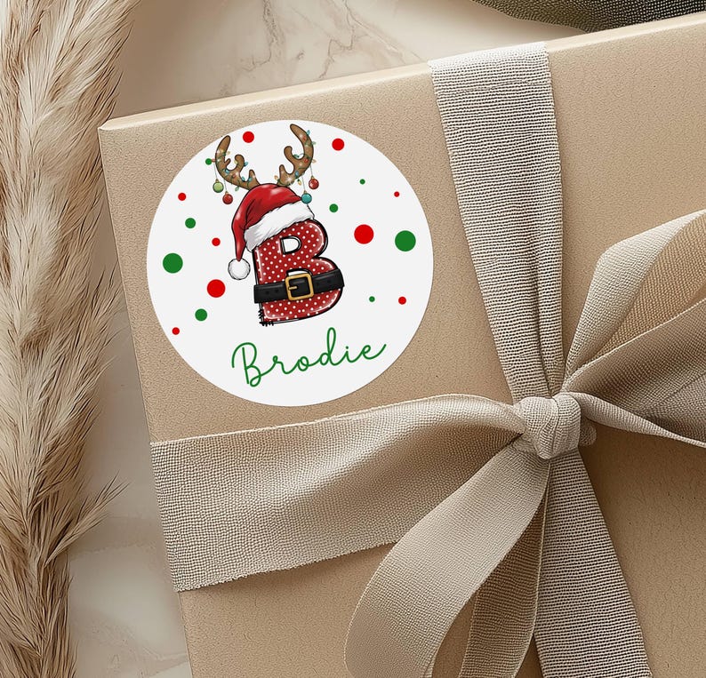 May include: A round white sticker with a Christmas theme, featuring the letter "B" in a Santa Claus outfit with reindeer antlers. The name "Brodie" is written in green script below the letter. Red and green dots surround the sticker.