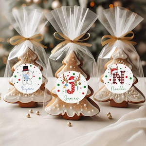 May include: Three Christmas tree-shaped gingerbread cookies, each individually wrapped in clear cellophane and tied with a gold ribbon. Each cookie has a round sticker with a festive design and a name: Ryan, James, and Noelle. The cookies are decorated with white icing.