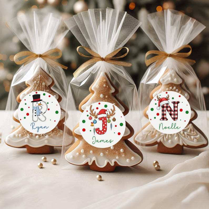May include: Three Christmas tree-shaped gingerbread cookies, each individually wrapped in clear cellophane and tied with a gold ribbon. Each cookie has a round sticker with a festive design and a name: Ryan, James, and Noelle. The cookies are decorated with white icing.