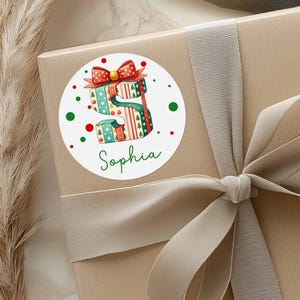 May include: A round sticker featuring a watercolor illustration of the letter "S" made of stacked gift boxes. The boxes are decorated with Christmas patterns and topped with a red bow. The name "Sophia" is written in green script below the letter.