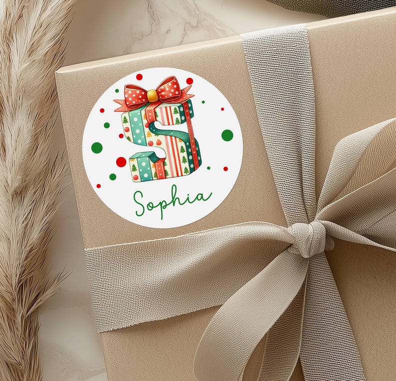 May include: A round sticker featuring a watercolor illustration of the letter "S" made of stacked gift boxes. The boxes are decorated with Christmas patterns and topped with a red bow. The name "Sophia" is written in green script below the letter.