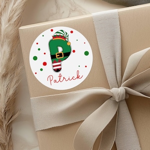 May include: A round sticker with the name "Patrick" in red script. The letter "P" is designed as an elf with a green hat and outfit. Red and green dots surround the sticker, which is placed on a gift box with a ribbon.