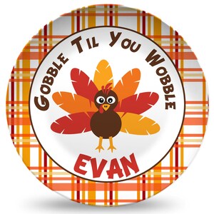 May include: A white ceramic plate with a red and orange plaid border. The center of the plate features a cartoon turkey with the text "Gobble Til You Wobble" and the name "Evan".