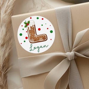 May include: A round sticker with a gingerbread letter "L" design, decorated with white icing, snowflakes, and holly. The name "Logan" is written in green script below the letter. Red and green dots surround the sticker, which is placed on a gift box.