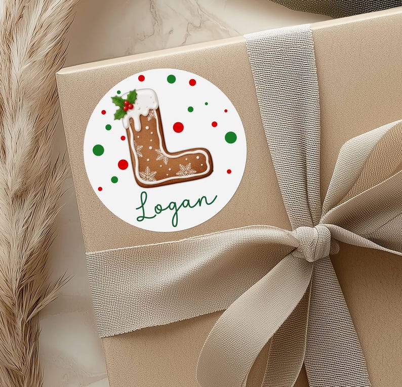 May include: A round sticker with a gingerbread letter "L" design, decorated with white icing, snowflakes, and holly. The name "Logan" is written in green script below the letter. Red and green dots surround the sticker, which is placed on a gift box.