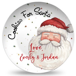 May include: White ceramic plate with a cartoon image of Santa Claus with a red hat and white beard. The plate has a green and gold snowflake pattern and the text "Cookies For Santa Love, Everly & Jordan".