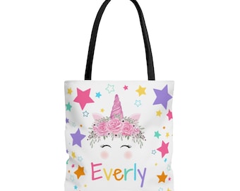 May include: White tote bag with a pink unicorn face with a flower crown and the name "Everly" printed in rainbow colors. The background is white with colorful stars.