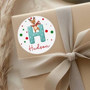 May include: A round sticker with a reindeer illustration on a gift box. The sticker features a teal letter "H" with a Christmas tree pattern, the name "Hudson" in red script, and red and green polka dots. The gift box is wrapped with a beige ribbon.