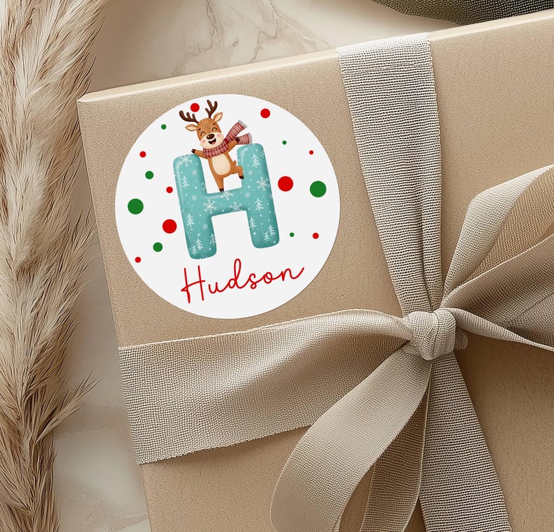 May include: A round sticker with a reindeer illustration on a gift box. The sticker features a teal letter "H" with a Christmas tree pattern, the name "Hudson" in red script, and red and green polka dots. The gift box is wrapped with a beige ribbon.