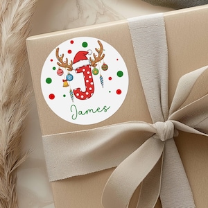 May include: A round white sticker with a Christmas theme, featuring a red letter "J" with white polka dots, antlers, and a Santa hat. The name "James" is written in green script below the letter. Red and green dots surround the design.