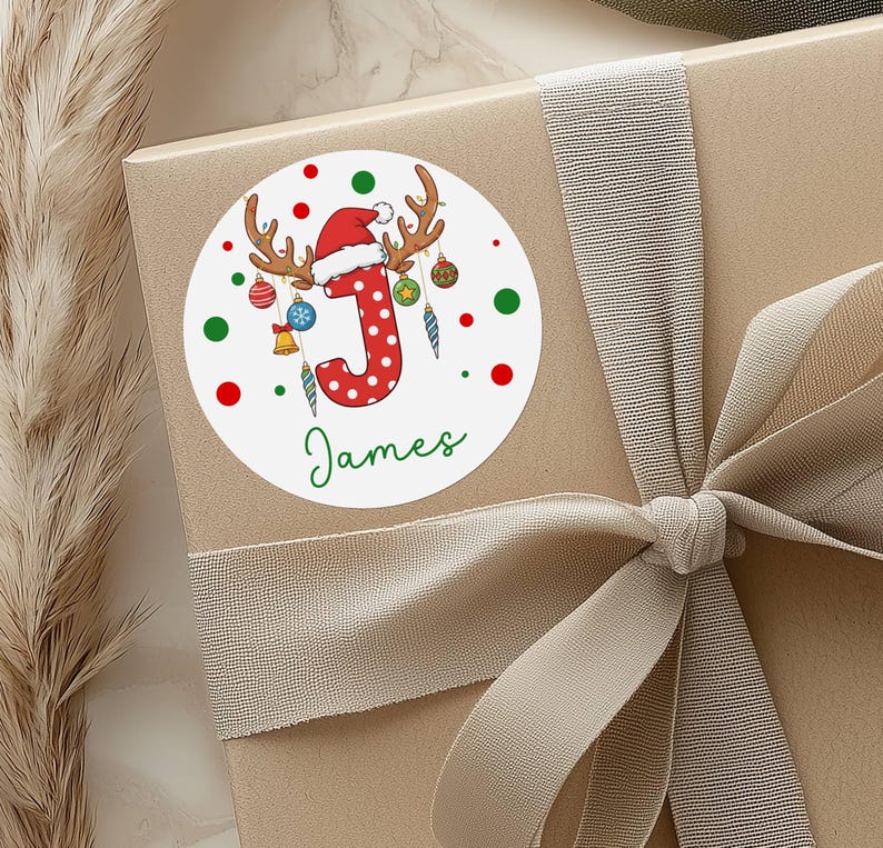 May include: A round white sticker with a Christmas theme, featuring a red letter "J" with white polka dots, antlers, and a Santa hat. The name "James" is written in green script below the letter. Red and green dots surround the design.