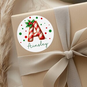 May include: A round Christmas sticker with the letter "A" in red and white candy cane stripes, surrounded by red and green polka dots. The name "Ainsley" is written in green script below the letter. The sticker is on a gift box with a ribbon.