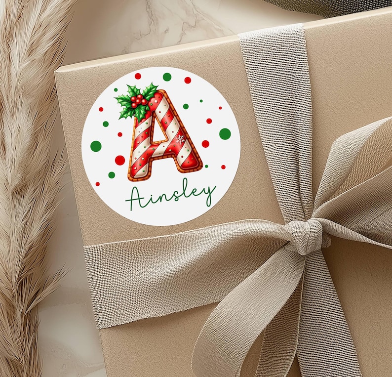 May include: A round Christmas sticker with the letter "A" in red and white candy cane stripes, surrounded by red and green polka dots. The name "Ainsley" is written in green script below the letter. The sticker is on a gift box with a ribbon.