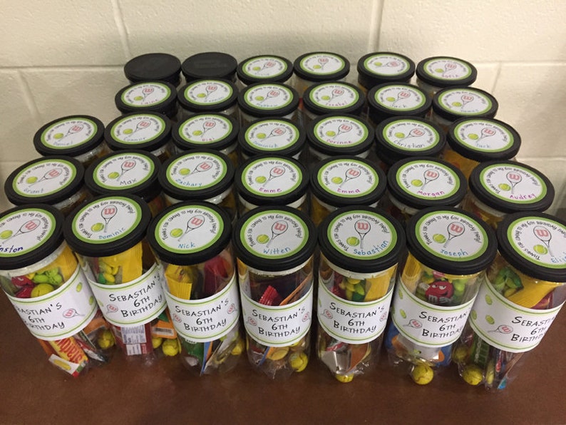 May include: A collection of clear plastic tubes filled with various small toys and candies, decorated with yellow and green labels that say "Sebastian's 6th Birthday". The tubes are arranged in rows on a wooden table.