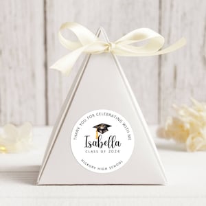 May include: White triangular favour box with a round sticker that says "Thank you for celebrating with me Isabella Class of 2024 Hickory High School" with a black graduation cap and tassel.