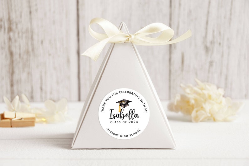 May include: White triangular favour box with a round sticker that says "Thank you for celebrating with me Isabella Class of 2024 Hickory High School" with a black graduation cap and tassel.