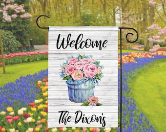 May include: Welcome to The Dixons' garden flag with a whitewashed wood background. A blue bucket filled with pink and white flowers is the centerpiece of the design.