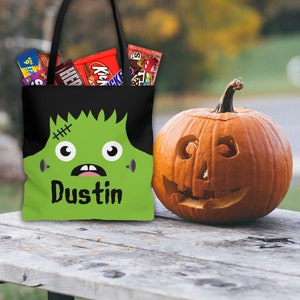 May include: A black and green Halloween trick-or-treat bag with a cartoon monster face and the name "Dustin" printed on it. The bag is filled with candy bars and is sitting on a wooden table next to a carved pumpkin.