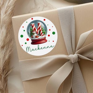May include: A round white sticker with a Christmas snow globe design. The snow globe features a snowman, candy cane trees, and the letter "M". The name "Makenna" is written in green script below the snow globe. Red and green dots surround the snow globe.
