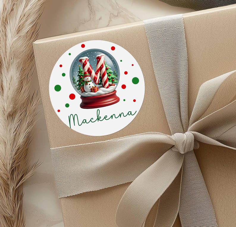 May include: A round white sticker with a Christmas snow globe design. The snow globe features a snowman, candy cane trees, and the letter "M". The name "Makenna" is written in green script below the snow globe. Red and green dots surround the snow globe.