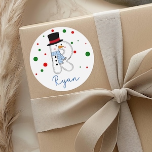 May include: A round sticker with a snowman design, a black top hat, and a blue scarf. The snowman is part of the letter "R" with the name "Ryan" in blue script below. Red and green polka dots surround the snowman on a white background.