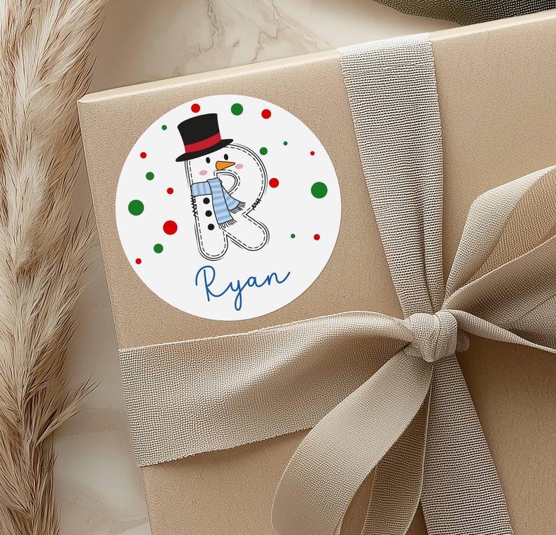May include: A round sticker with a snowman design, a black top hat, and a blue scarf. The snowman is part of the letter "R" with the name "Ryan" in blue script below. Red and green polka dots surround the snowman on a white background.