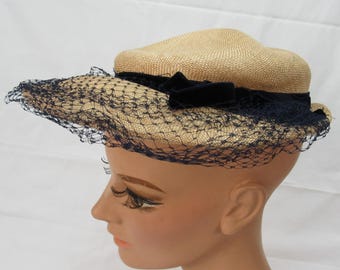 Puede incluir: Sombrero de paja vintage con copa redondeada y ala ancha. El sombrero presenta una cinta de terciopelo azul marino oscuro y un velo de red a juego. El sombrero es de color beige claro y se exhibe en una cabeza de maniquí.