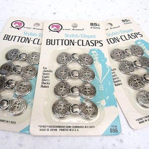May include: Three identical retail cards, each containing four sets of silver-toned metal button-clasps. The packaging features the text "Stylish/Elegant BUTTON-CLASPS" and "85c 4 Sets." The metal clasps have a decorative, ornate circular design.