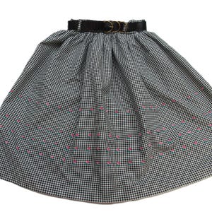 Peut inclure: Jupe à carreaux noirs et blancs avec une ceinture noire et une boucle dorée. La jupe présente des motifs floraux brodés roses et verts près de l'ourlet. Style classique.