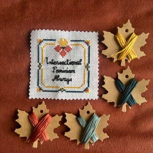 Op de afbeelding: Een witte kruissteek sampler met de tekst "Intersectional Feminism Always" in een zwart frame. Vier bruine, esdoornblaadjesvormige stukken stof met borduurgaren in verschillende kleuren zijn inbegrepen.