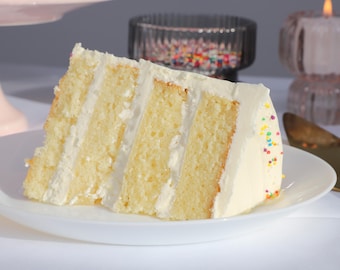 May include: A slice of layered cake with white frosting and colorful sprinkles on a white plate. The cake has a light yellow sponge and is decorated with a gold knife and a lit candle in the background. The cake is a dessert.