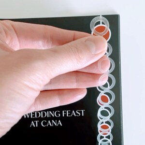 May include: A black book with the title "Wedding Feast at Cana" on the cover. A clear plastic bookmark with a repeating circle design is attached to the spine of the book.