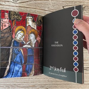May include: An open book with a stained glass image of two women on the left page. The right page has the text "THE VISITATION" and "2nd Joyful for greater love of neighbor". The spine of the book has a vertical row of alternating red and blue circles.
