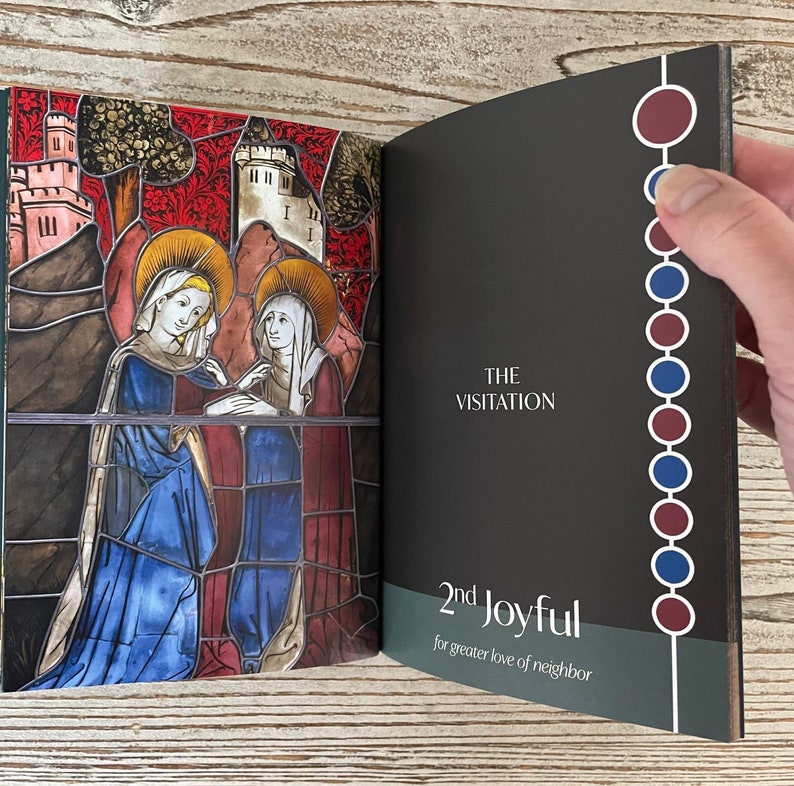 May include: An open book with a stained glass image of two women on the left page. The right page has the text "THE VISITATION" and "2nd Joyful for greater love of neighbor". The spine of the book has a vertical row of alternating red and blue circles.