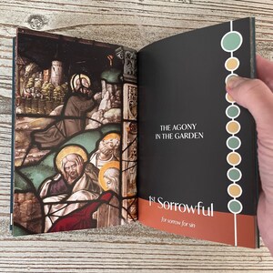 May include: An open book with a stained glass image of Jesus praying in the garden of Gethsemane. The text on the right page reads "The Agony in the Garden" and "1st Sorrowful for sorrow for sin".