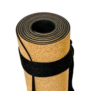 May include: A tightly rolled exercise mat featuring a tan, cork-like textured outer surface and a solid black inner layer. The mat is secured with a wide, black woven fabric strap wrapped around the center, creating a cylindrical shape.