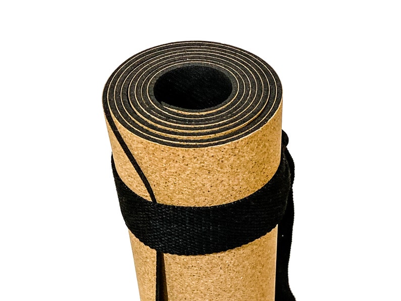 May include: A tightly rolled exercise mat featuring a tan, cork-like textured outer surface and a solid black inner layer. The mat is secured with a wide, black woven fabric strap wrapped around the center, creating a cylindrical shape.