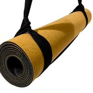 May include: A rolled yoga mat with a tan cork-textured top surface and a black rubberized base. The mat is secured by a black woven fabric carrying strap that loops around both ends of the roll, allowing it to be suspended for transport or storage.
