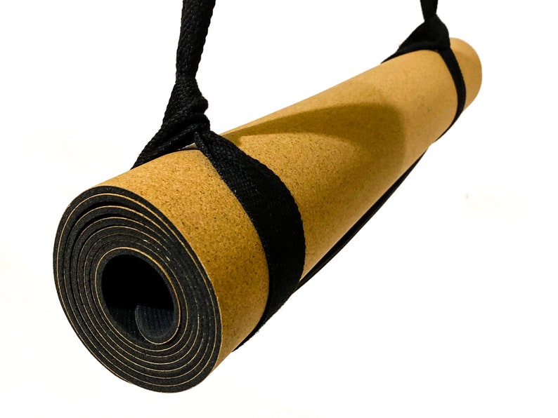 May include: A rolled yoga mat with a tan cork-textured top surface and a black rubberized base. The mat is secured by a black woven fabric carrying strap that loops around both ends of the roll, allowing it to be suspended for transport or storage.