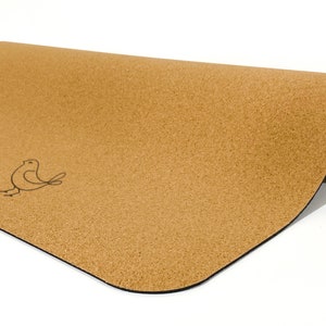 May include: A tan cork yoga mat partially rolled up, featuring a smooth, speckled texture and a simple black line drawing of a small bird on the left side. The mat has rounded corners and a dark, solid black base layer visible on the underside.