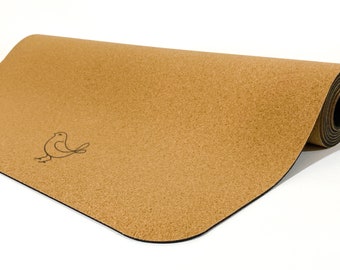 Op de afbeelding: Een gedeeltelijk opgerolde yogamat van beige kurk met een gladde, gespikkelde textuur en een eenvoudige zwarte lijntekening van een kleine vogel aan de linkerkant. De mat heeft afgeronde hoeken en een donkere, effen zwarte onderlaag.