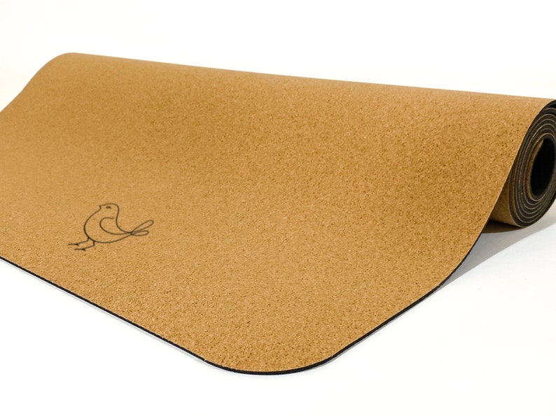 May include: A tan cork yoga mat partially rolled up, featuring a smooth, speckled texture and a simple black line drawing of a small bird on the left side. The mat has rounded corners and a dark, solid black base layer visible on the underside.