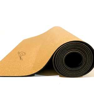 May include: A partially rolled yoga mat featuring a light tan, textured cork top surface and a solid black rubberized base. A small, simple black line drawing of a bird is printed on the flat portion of the cork surface near the center.
